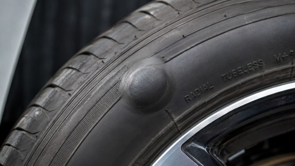 A close-up view of a car tire sidewall displaying a bulge, indicating internal structural damage from a pothole or curb impact.